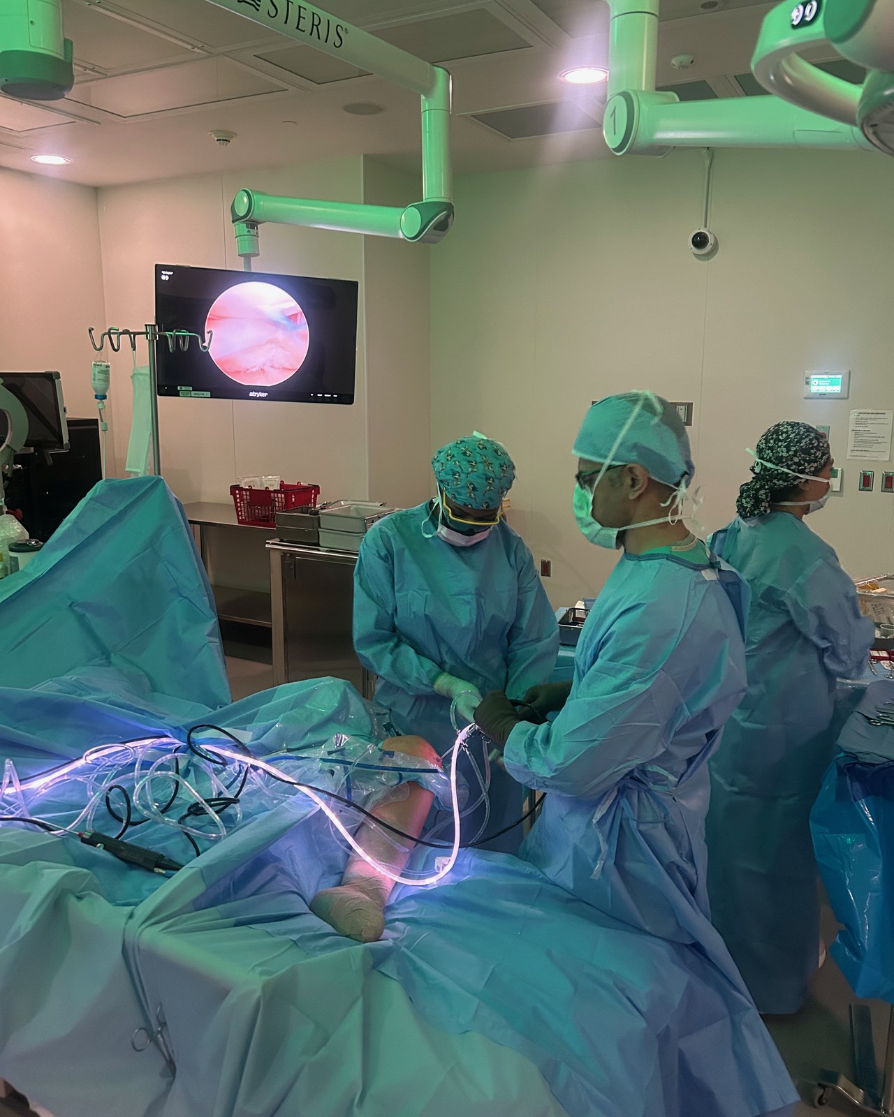 Dr. Dewan performing knee arthroscopy in the operating room, with the arthroscope's view of the inside of the knee joint displayed on the monitor
